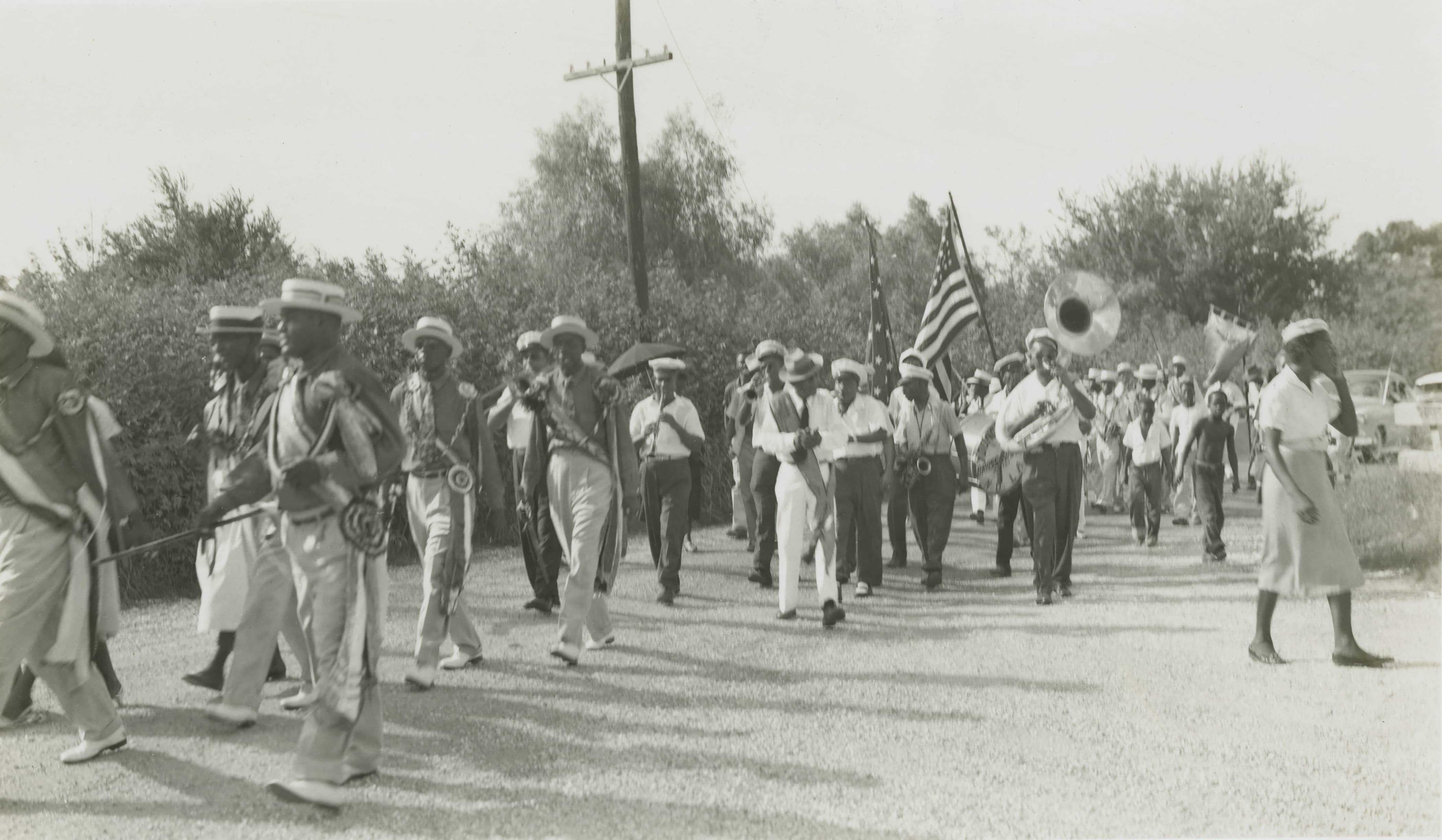A black and white photo of a marching band parading down a rural road. Band members play instruments like drums and a tuba, while carrying an American flag. Spectators walk alongside, and trees line the background.