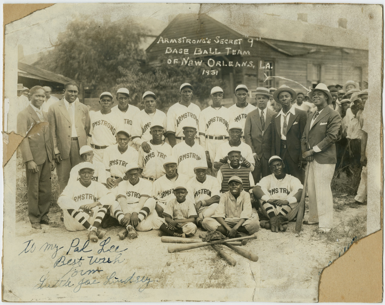 Black and white photograph of Armstrongs Secret 9 Baseball Team from New Orleans, 1931. The players, wearing uniforms with Armstrong on them, are posed with bats, alongside coaches or supporters in suits and hats. Handwritten dedication at the bottom.
