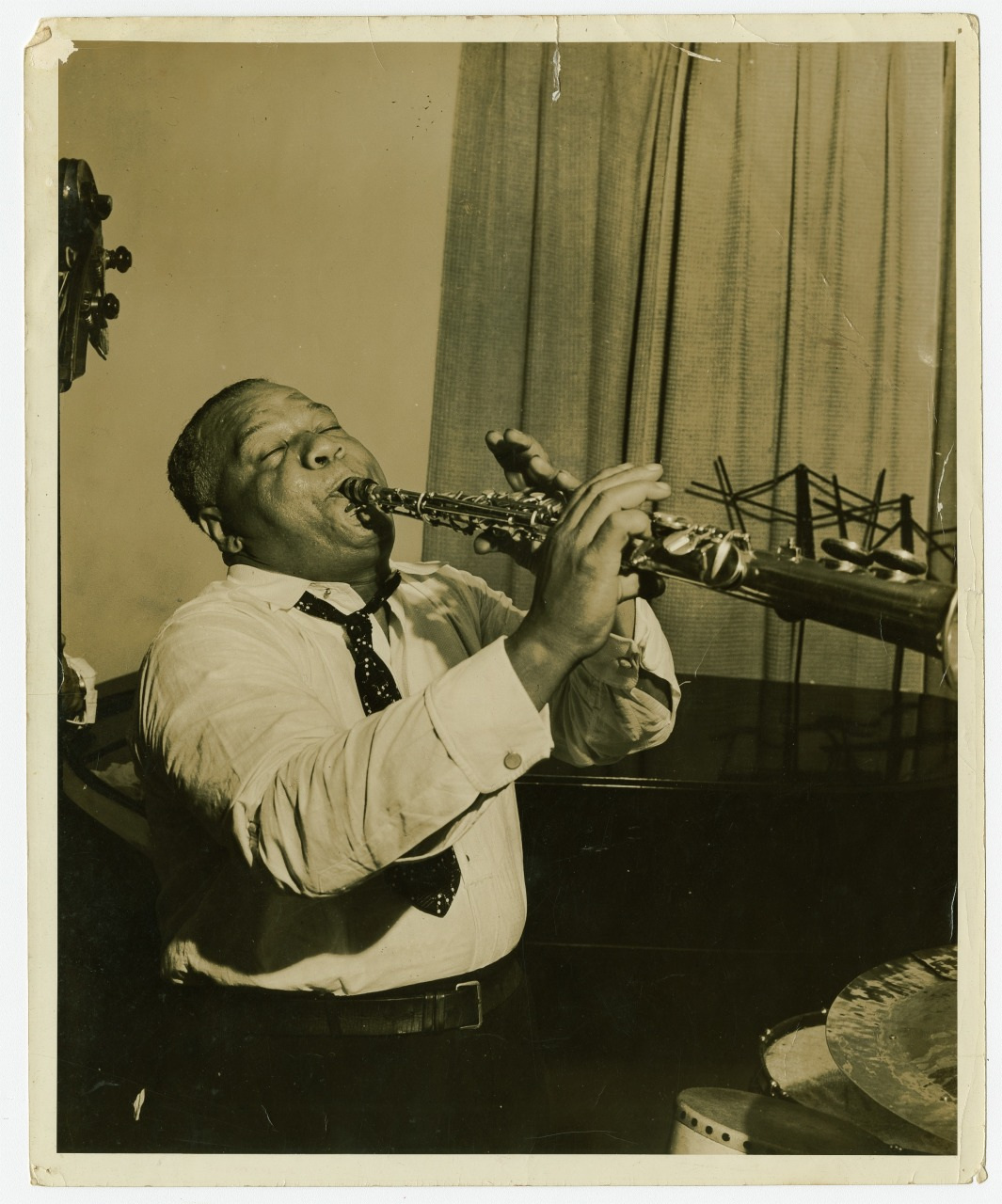 A musician passionately plays a saxophone in a room with curtains. He is wearing a light shirt and a dark tie. A double bass and music stand are visible in the background.