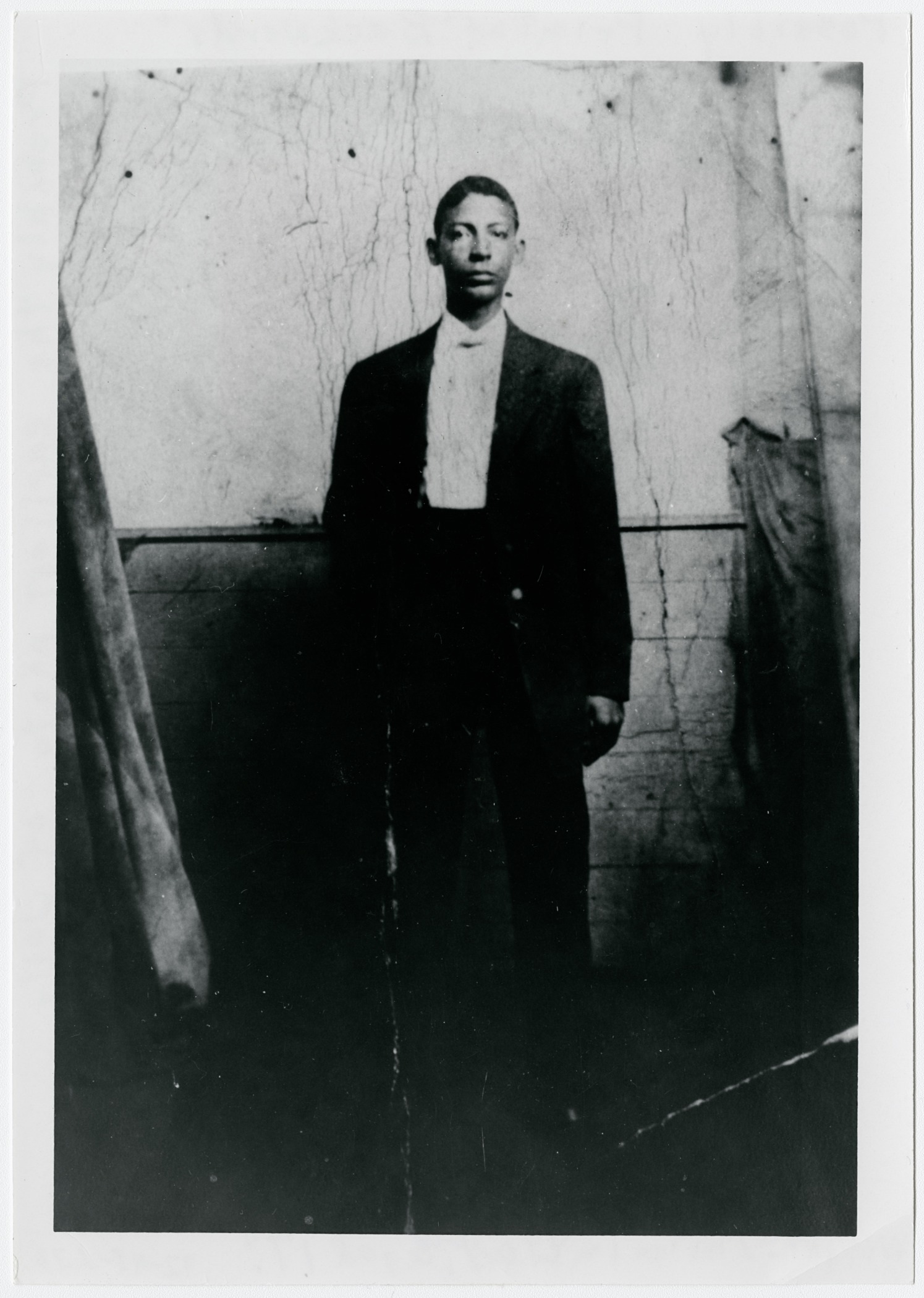 A historical black and white photo of a man standing against a simple backdrop. The man is wearing a dark suit and a light shirt, with his hand at his side. The setting appears worn, adding a vintage feel to the image.