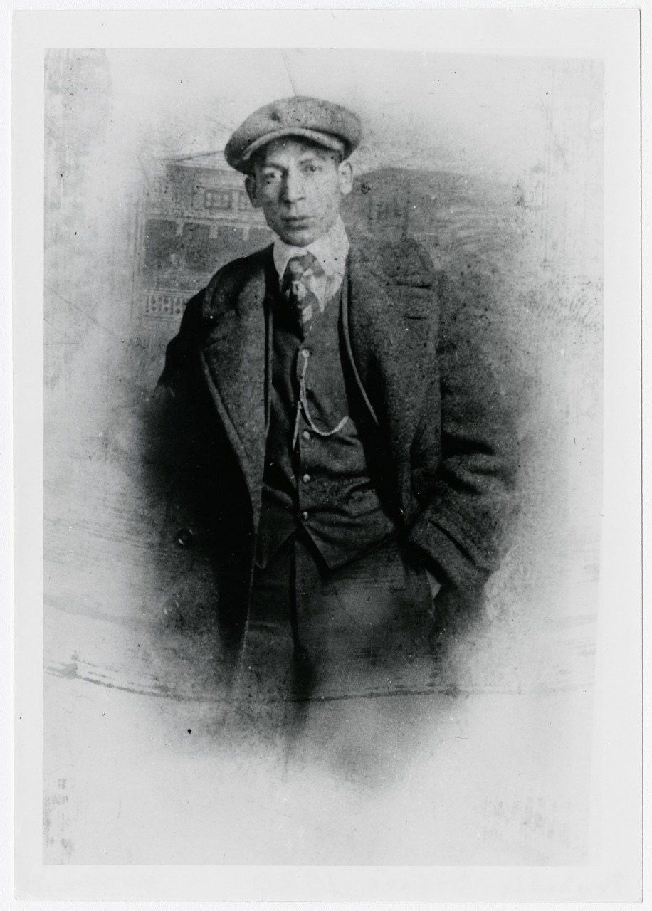 A vintage black and white photograph of a man wearing a flat cap, suit, tie, and overcoat. He stands in a relaxed pose with one hand in his pocket. The background is faded, giving the image an antique, nostalgic feel.