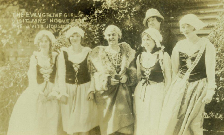A group of six women poses outdoors, dressed in period costumes with bonnets and lace-up bodices. One woman is wearing a fur-trimmed coat. Text on the image reads, The Evangeline Girls visit Mrs. Hoover at the White House.