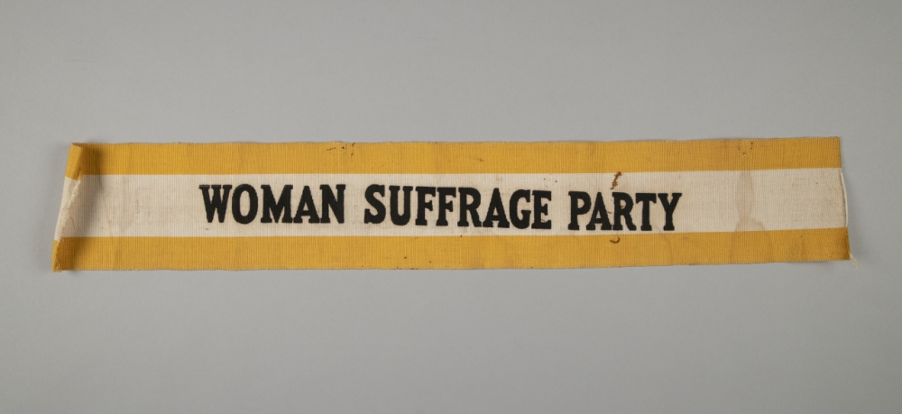 A vintage banner with a yellow border and white center, displaying the text WOMAN SUFFRAGE PARTY in bold black letters. The background is plain and light-colored.