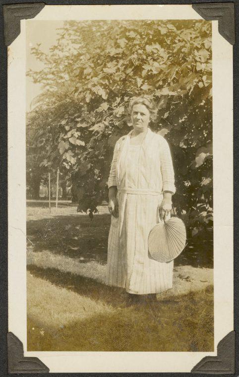 A vintage photograph of a woman standing outdoors in front of a large bush. She wears a long dress and holds a hand fan. The photo has a sepia tone, evoking an early 20th-century style.