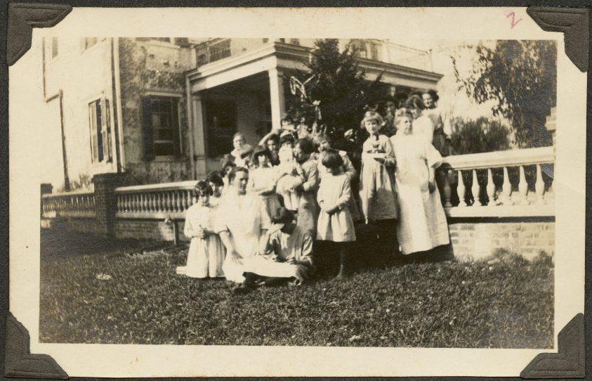 A vintage photo of a group of women and children gathered in front of a large house with a porch. Some are seated while others stand on the grass, wearing period clothing. The scene appears candid and informal.