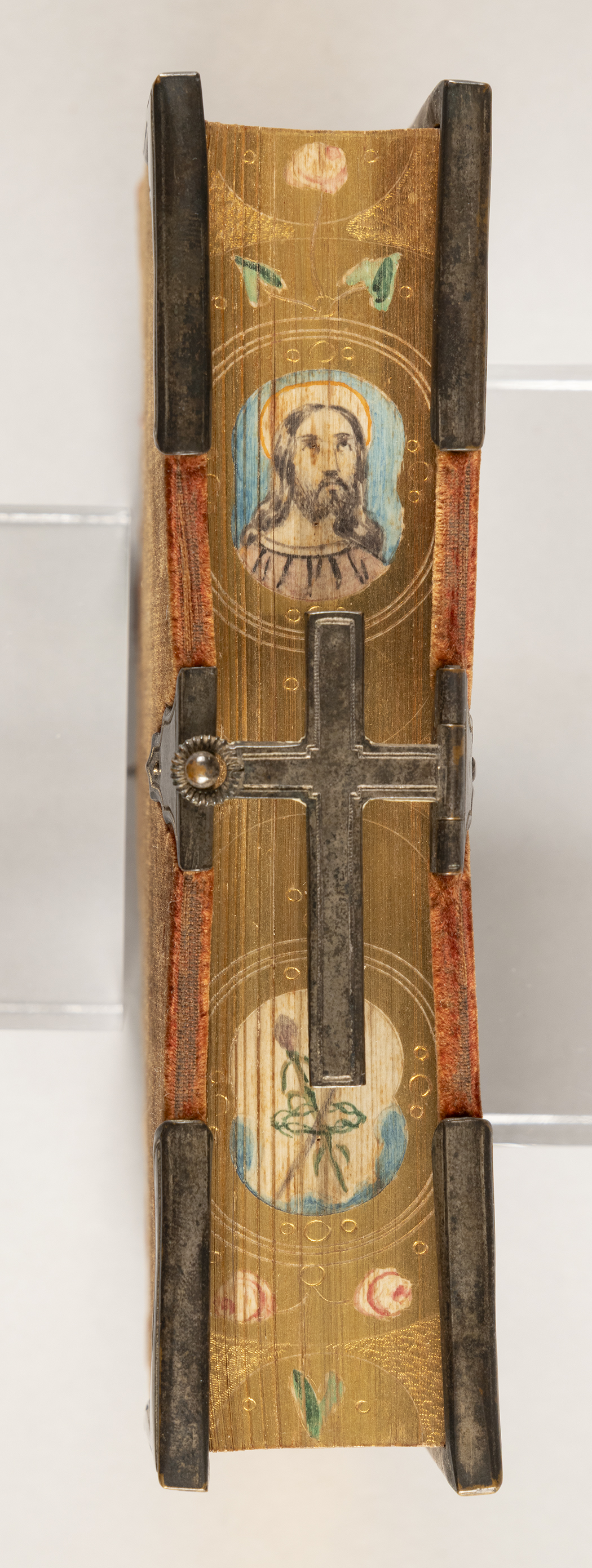 A profile view of a red belvet-bound prayer book, showing the painted edge of the pages which shows a depiction of Jesus. The clasp enjoining the front and back cover is a metal cross.