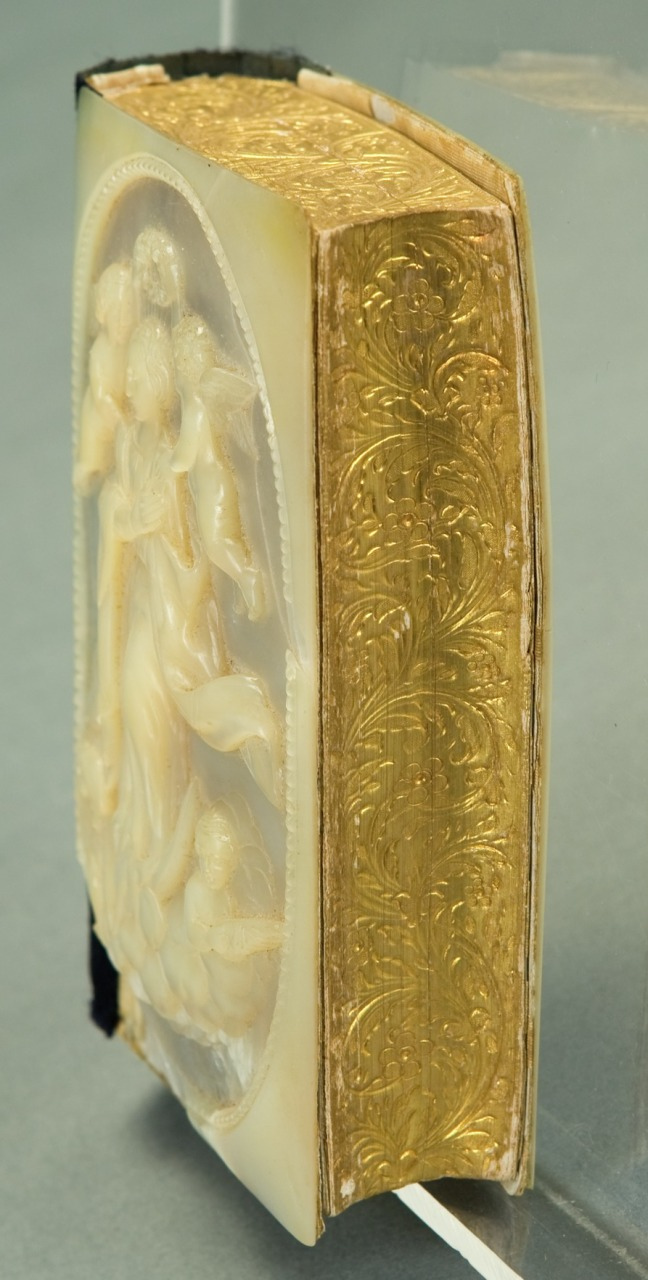An intricately sculpted, oval-shaped mother-of-pearl relief adorns the cover of a gold-embossed book. The relief depicts a group of angels. The books spine features ornate, swirling gold designs.