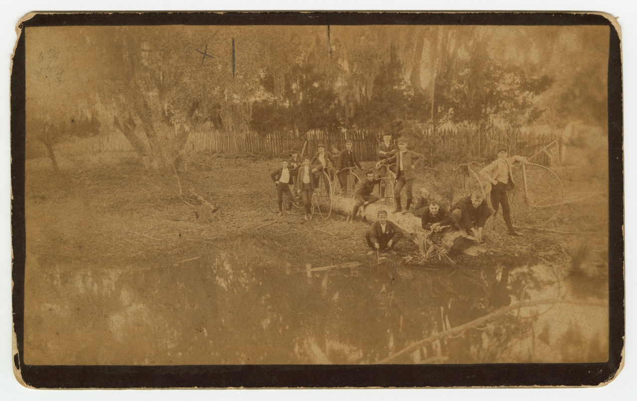 Sepia-toned photo of a group of people standing and sitting near a tree by a pond. Some are leaning on the tree, and a few are sitting on its trunk. A wooden fence and trees are visible in the background.