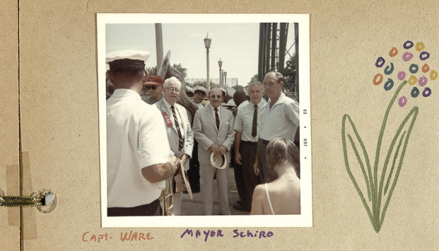 A group of men in formal attire stand outdoors, surrounded by others. One man wears a military-style cap. They appear to be at a public event. There is a hand-drawn flower on the side of the photo.