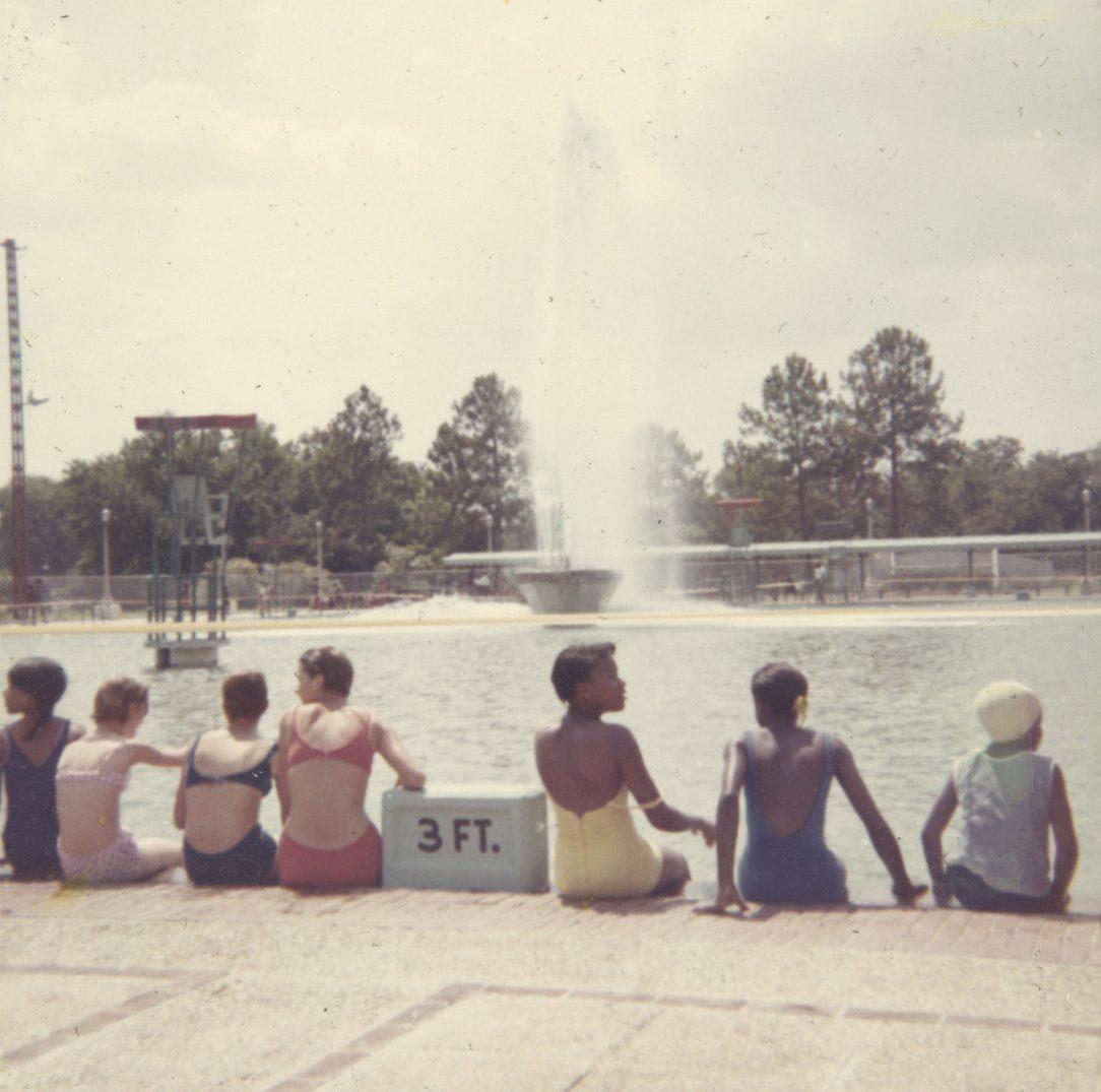 A group of children in swimsuits sit by the edge of a large outdoor pool, with a sign reading 3 FT. A fountain sprays water in the center of the pool, and trees are visible in the background under a partly cloudy sky.
