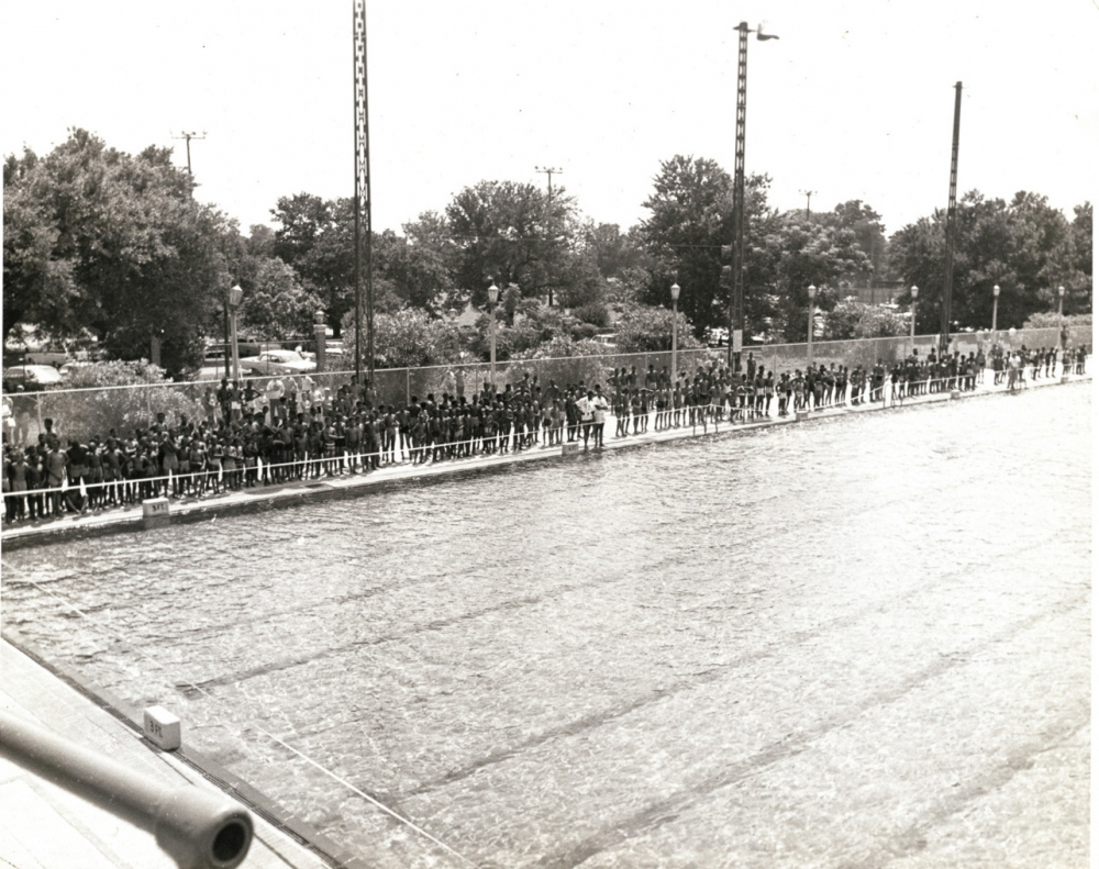 A large crowd of people stands along the edge of an outdoor swimming pool, separated by a fence. Trees and parked cars are visible in the background. The image is in black and white, suggesting it is from a past era.