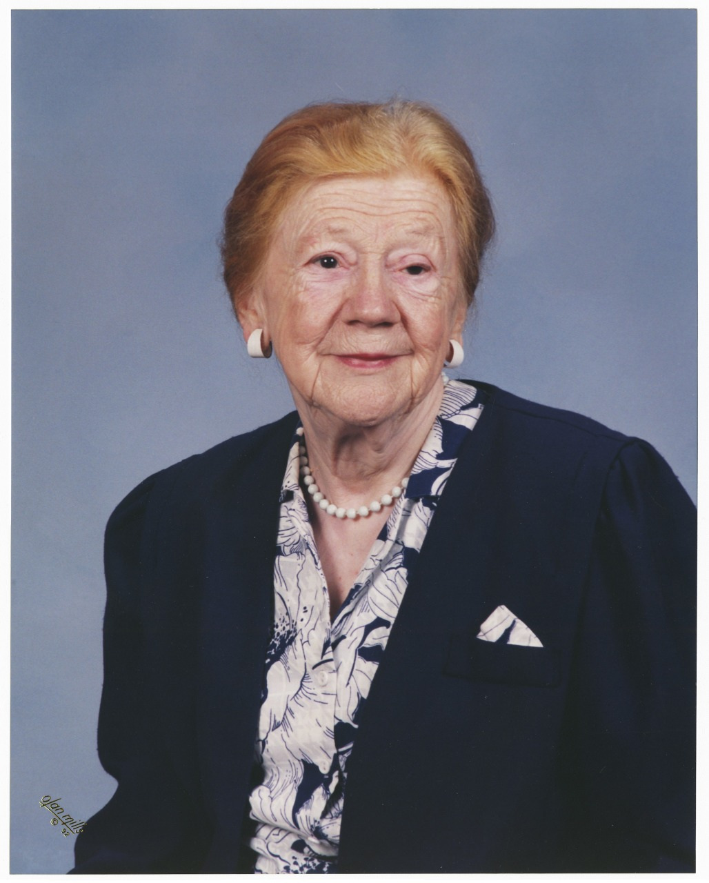 An elderly woman with short, light brown hair is dressed in a navy blazer over a patterned blouse. She is wearing a pearl necklace and white earrings, and she smiles softly against a plain blue backdrop.
