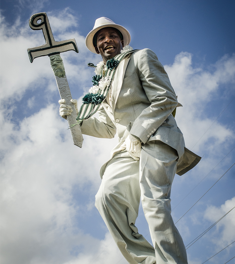 A man dressed in an elegant white suit, white gloves, and a matching fedora smiles confidently while holding a decorated prop depicting the number nine. He is elevated above the crowd, against a backdrop of a bright blue sky and scattered clouds.