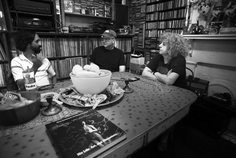 A black and white photo of authors Michael Tisserand and Allison Fensterstock sitting at a table talking to Jude Matthews. A copy of the book "Bunny Matthews: His Life, Art, and Obsessions" is on the table.