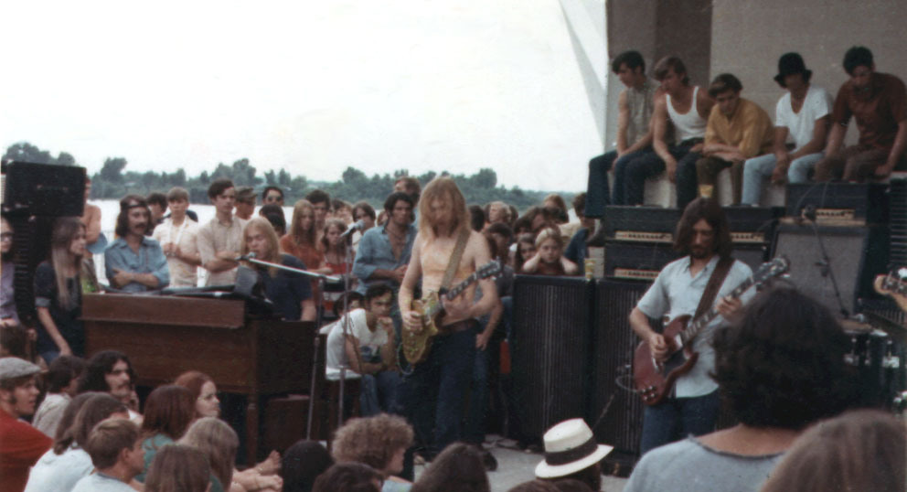 A classic rock band performs on an outdoor stage. Two guitarists and a keyboard player are visible, surrounded by a large crowd of spectators sitting and standing. The audience is engaged, enjoying the music in a casual, sunny setting.