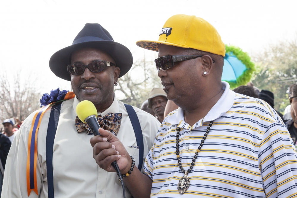 Two men in hats are speaking at an outdoor event. One wears a suit with a bow tie and is holding a microphone, while the other wears sunglasses, a striped shirt, and a yellow cap. A crowd is visible in the background.