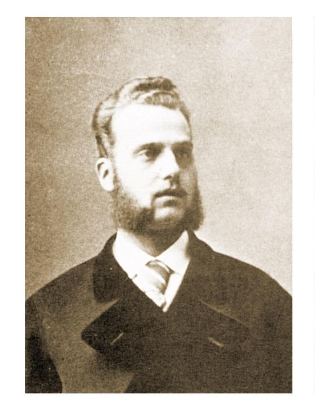 Vintage sepia-toned portrait of a man with a beard and sideburns. He wears a dark suit, a white shirt, and a tie, looking slightly to the side. The background is plain, emphasizing the formal attire and hairstyle typical of an earlier era.