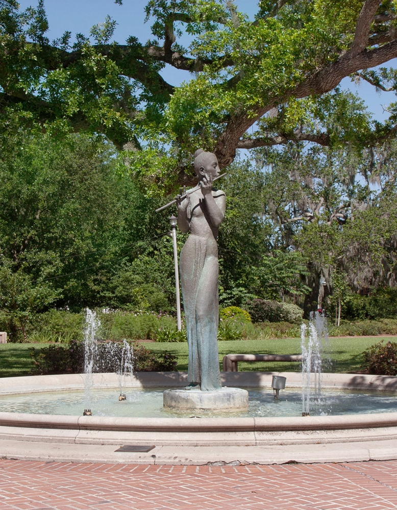 A statue of a person playing the flute stands in the center of a circular fountain surrounded by small water jets. Lush green trees and grass are visible in the background, creating a serene park setting.