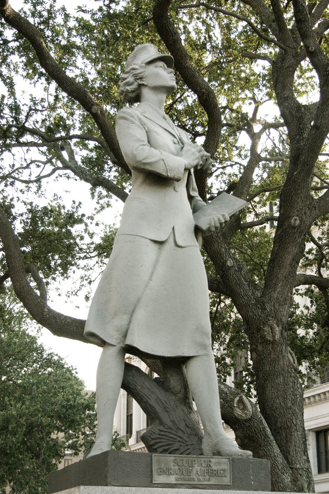 A stone statue of a woman in a suit holding a book, standing confidently with one leg propped on a rock. The sculpture is set against a backdrop of leafy trees and a building.