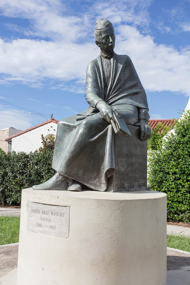 A statue of Sophie Bell Wright, a teacher from 1866 to 1912, sits on a pedestal. The sculpture depicts a seated figure holding a book, with a shawl draped over her shoulders. The background features greenery and a partly cloudy sky.