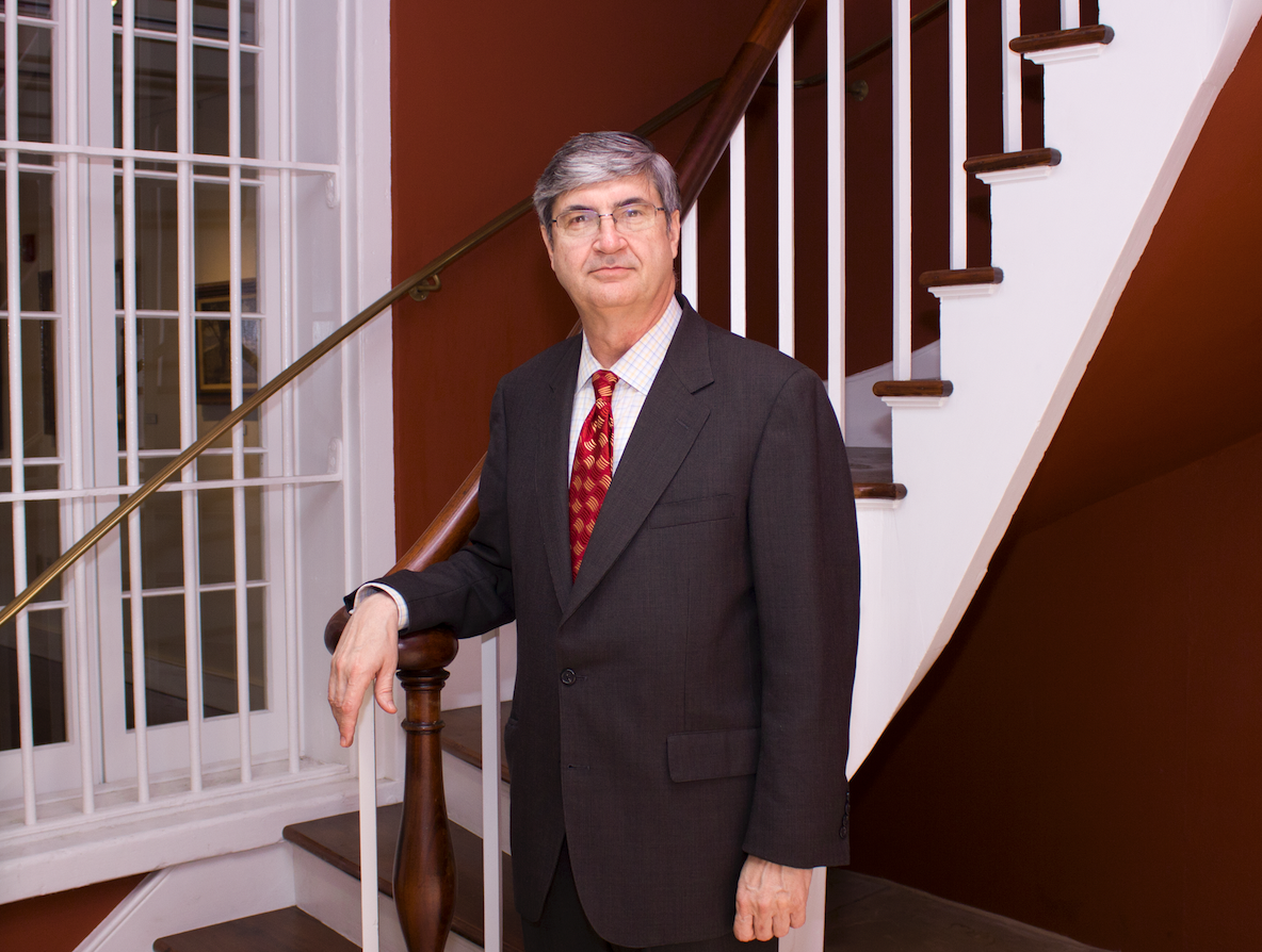 A man in a dark suit and patterned red tie stands on a staircase with white railings, in front of a large window with bars. The staircase is set against a red wall. He appears to be posing for a formal photo.