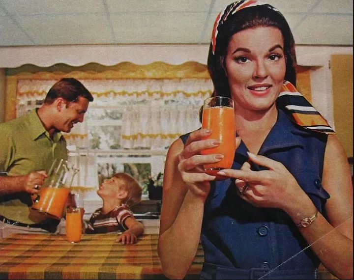 A screenshot from a commercial featuring Anita Bryan, who was the spokesperson for the Florida Citrus Commission. She points at a glass of orange juice with characters protraying her husband and child in the background, which looks like a typical kitchen of the era.