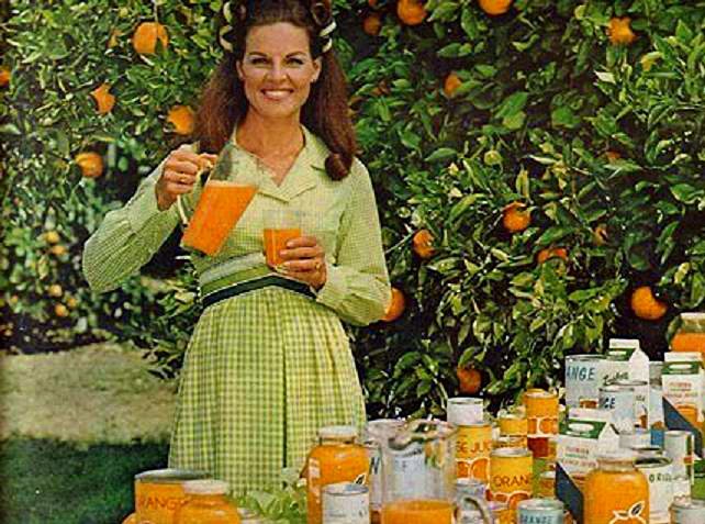 A screenshot from a commercial featuring Anita Bryan, who was the spokesperson for the Florida Citrus Commission. She is wearing a traditional-looking checkered dress while pouring orange juice into a glass, while standing in front of an orange grove next to a table of orange juice products.
