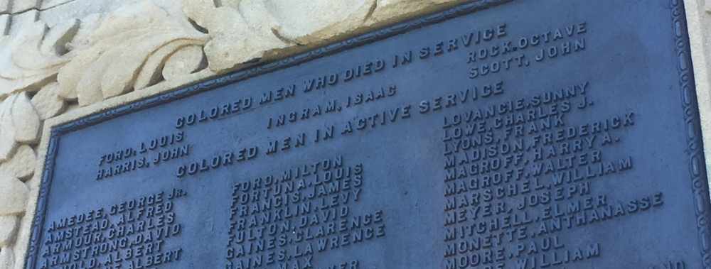 A memorial plaque on a stone wall honors Colored Men Who Died in Service. The names of several individuals are inscribed on the plaque, surrounded by decorative carvings.