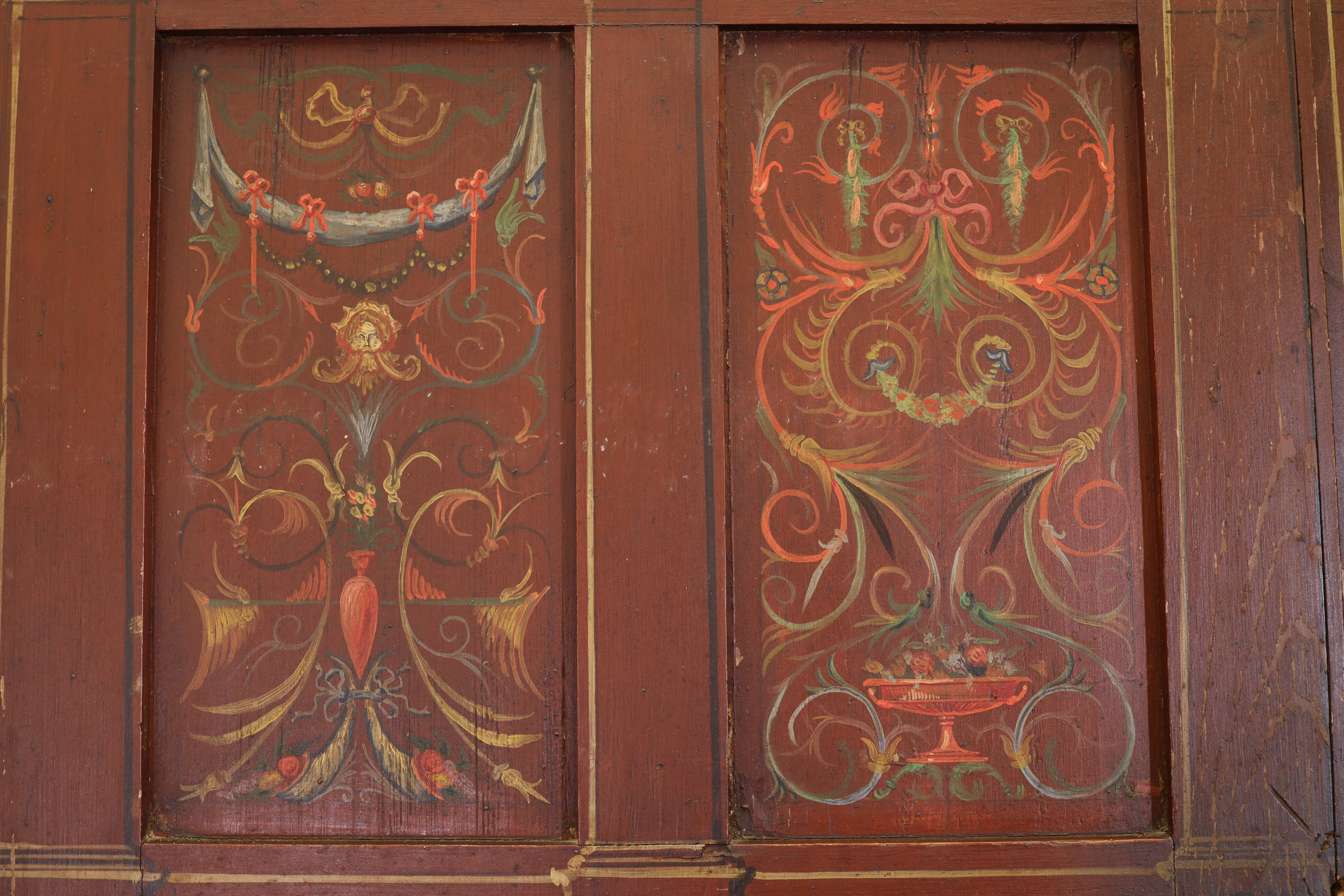 An intricately painted antique wooden panel featuring symmetrical ornate designs. The left side has floral motifs, ribbons, and a face, while the right shows floral patterns with a central urn. The colors are primarily red, green, and gold.