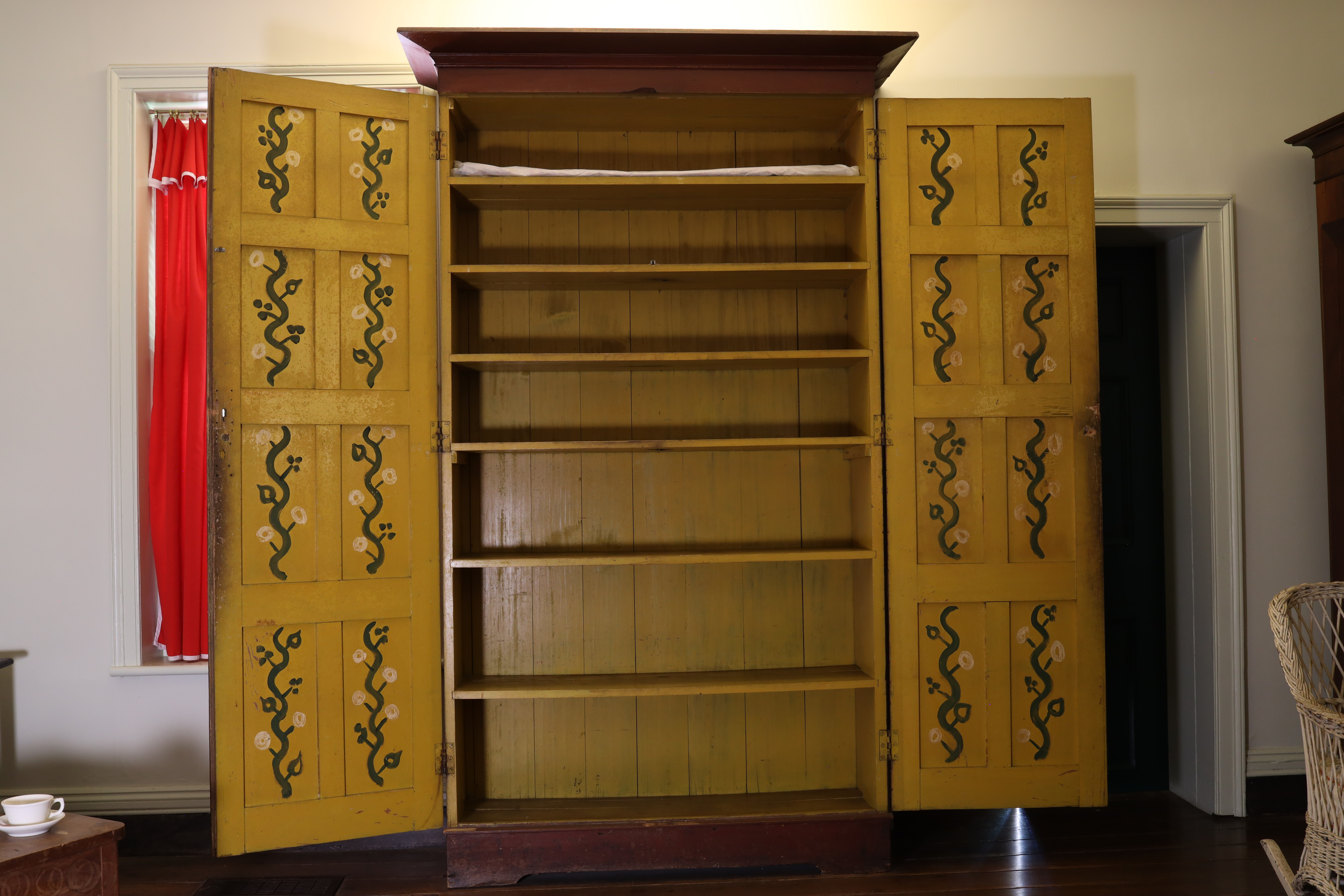 An open, vintage wooden cabinet with ornate yellow doors displaying floral patterns. The interior has multiple empty shelves. A red curtain is partially visible to the left. The room is warmly lit.