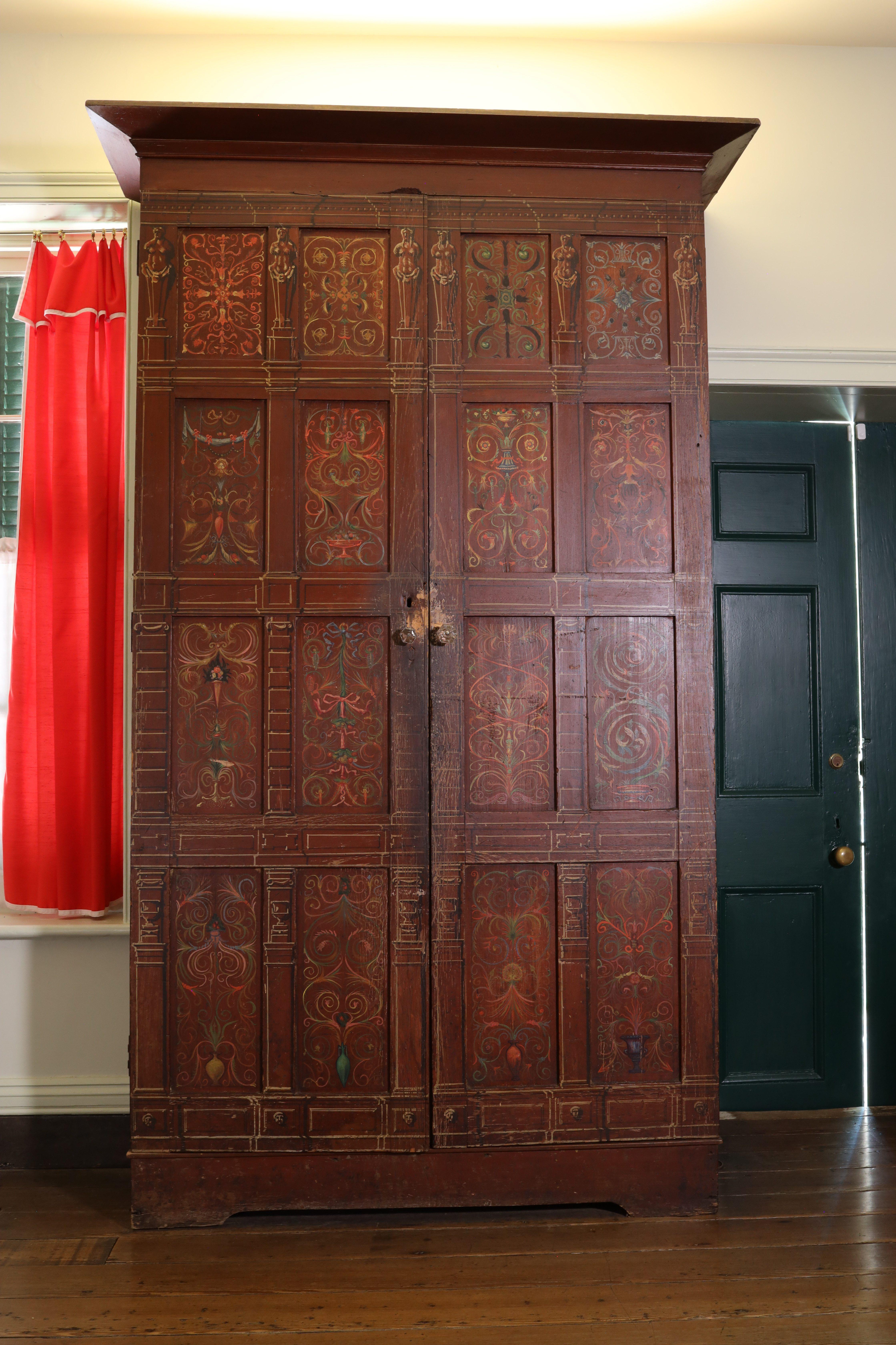 A tall, ornate wooden wardrobe with intricate carvings and patterns stands against a wall. It has two doors with metal handles. To the left is a window with red curtains, and to the right is a closed green door.