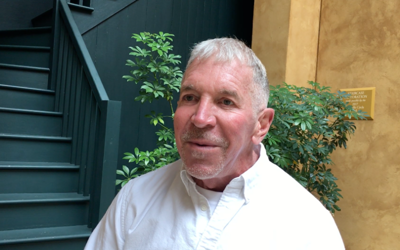 A man with short gray hair and a white button-up shirt stands in front of a staircase and green plants. The setting has a warm, inviting atmosphere with a mix of natural and modern elements.