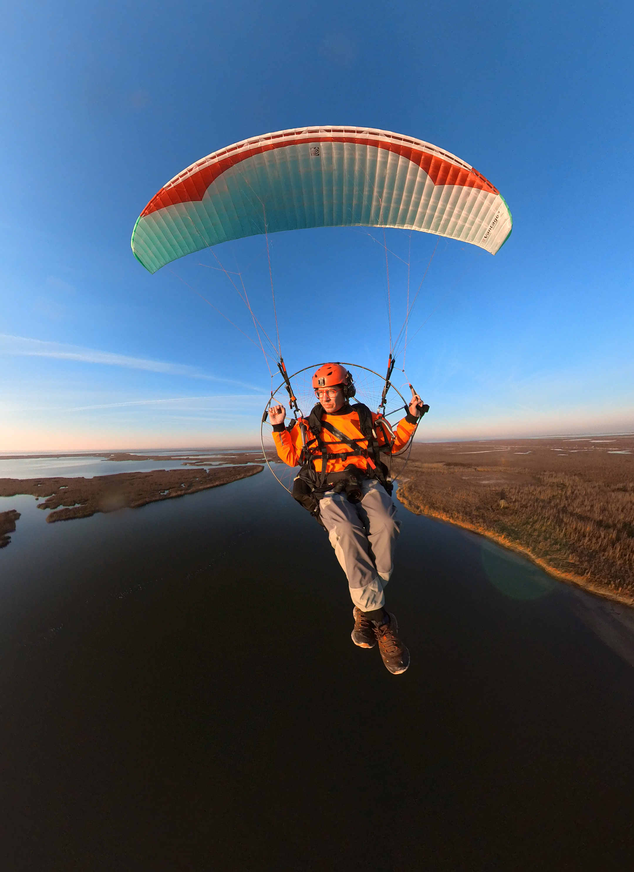 Photographer Ben Depp flies his paraglider with Louisiana marshland extending into the distance behind him. He wears an orange outfit and helmet.