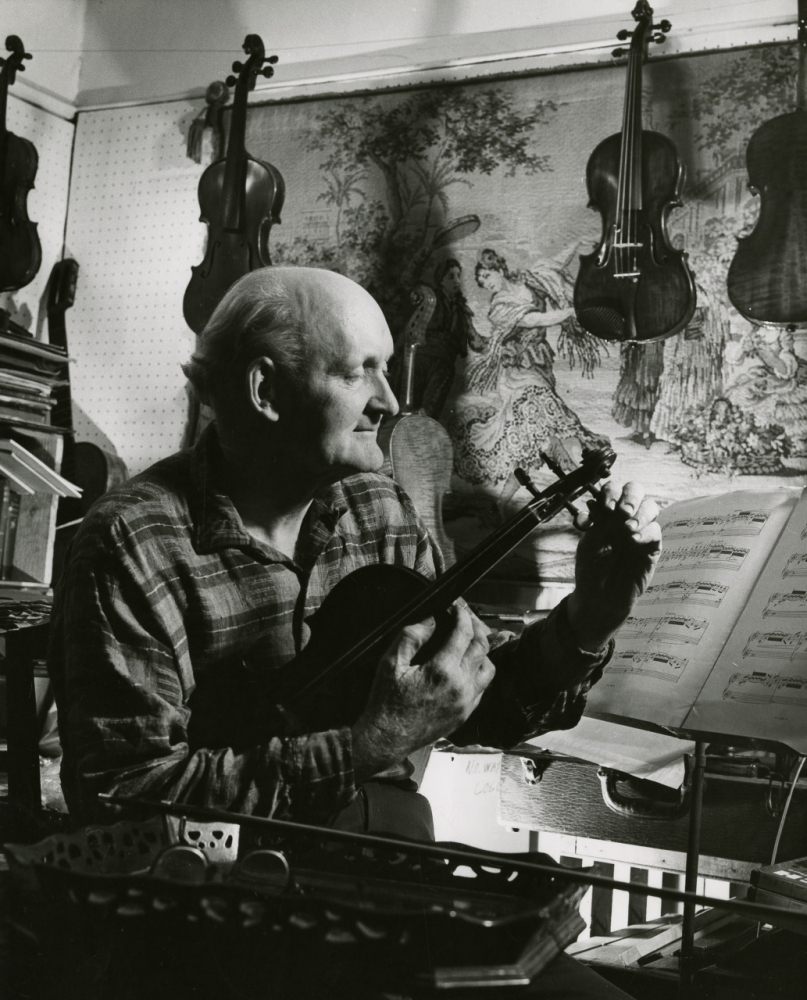 An elderly man sits in a room surrounded by violins, adjusting the strings of a violin in his hands. Sheet music and a tapestry depicting a classical scene are visible in the background. The setting is dimly lit, creating a serene and focused atmosphere.