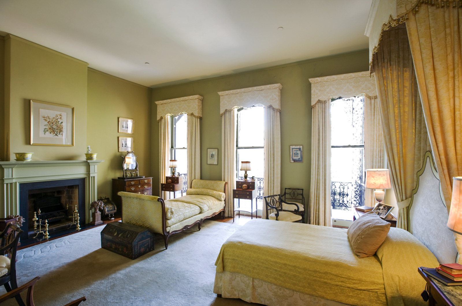 A spacious, elegant bedroom with pale yellow walls features a classic fireplace, a large bed, a chaise lounge, and multiple windows draped with lace curtains, allowing soft light to illuminate the room. Vintage furniture and decor complete the setting.
