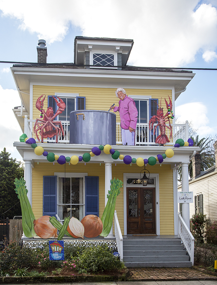 A yellow house decorated with a large cutout of a person standing by a giant pot, surrounded by oversized lobsters and vegetables. Beads and Mardi Gras symbols adorn the front, enhancing the festive atmosphere.