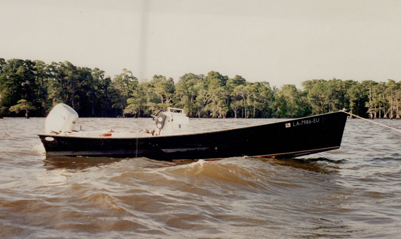 A small black fishing boat with LA 7366 EU on the side floats on a river. It has an outboard motor and is surrounded by trees along the riverbank. The water appears slightly choppy.