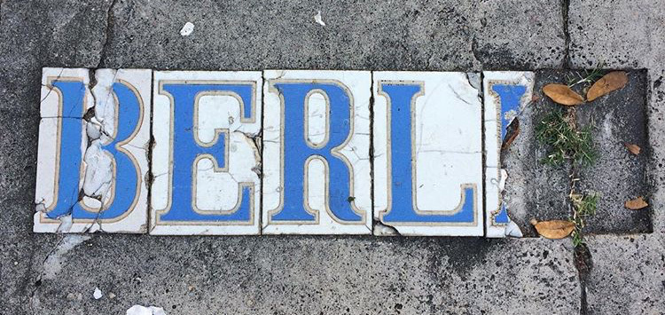Tiles on a sidewalk spell out BERL with cracked blue letters on a white background. The surrounding area is worn, with grass and dry leaves on the right.