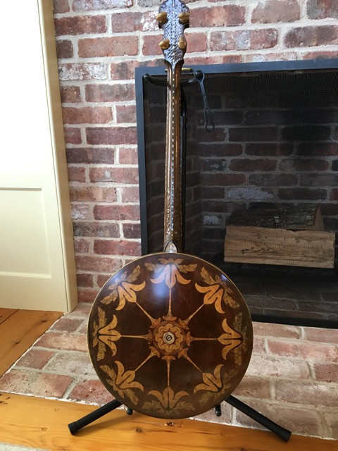 A vintage banjo with an ornate, intricate floral design on its back rests on a stand. The background features a brick fireplace and wooden floor, creating a cozy setting.