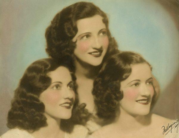 A vintage portrait of three women with wavy dark hair and bright smiles. They are dressed in light-colored clothes and have rosy cheeks. They are styled in an elegant, classic manner against a softly blurred backdrop.