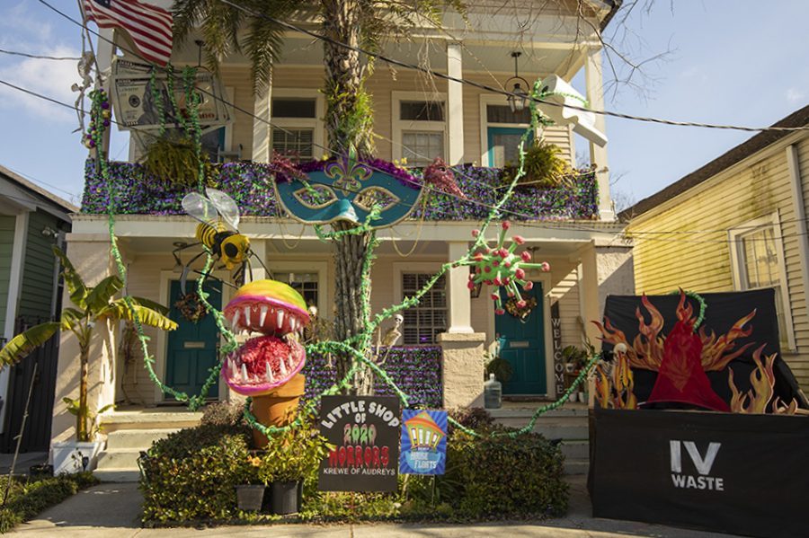 A two-story house is decorated with Mardi Gras-themed elements, including a large potted plant with a pink open mouth, colorful beads, a giant bee, and a sign reading Little Shop 2020 Krewe of House Floats. The porch has greenery and hanging decor.