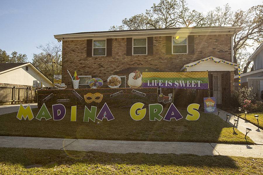 A brick house decorated for a Mardi Gras celebration with a banner reading LIFE IS SWEET and colorful lawn signs spelling MADINA GRAS. Theres also a mask and images of food and drinks in the background.