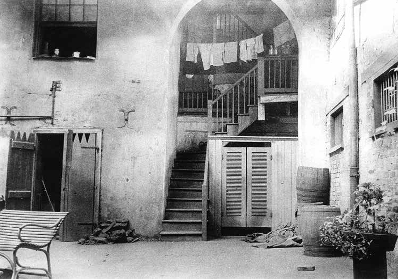 A vintage black and white photo of a courtyard with stairs leading to a second floor. Clothes hang on a line above, and there is a bench, a barrel, a potted plant, and an open door on the ground level. The walls and windows appear worn.