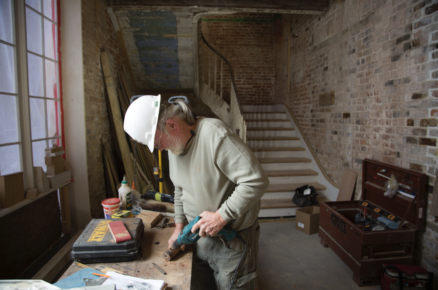 An older man wearing a white hard hat works on a construction project inside a rustic building. He is standing at a wooden workbench using a power tool. The space has exposed brick walls and a staircase in the background. Tools are scattered on the bench.