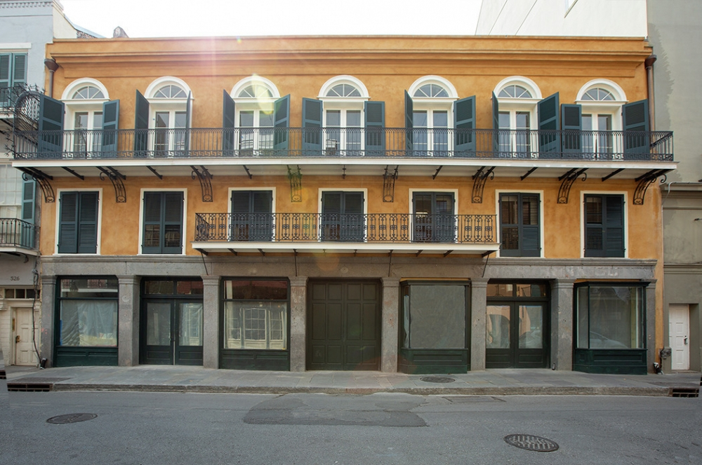 A two-story building with a yellow facade and black shutters, featuring large arched windows and balconies. Sunlight creates a lens flare effect across the upper part. The ground floor has covered storefronts and a central entrance.