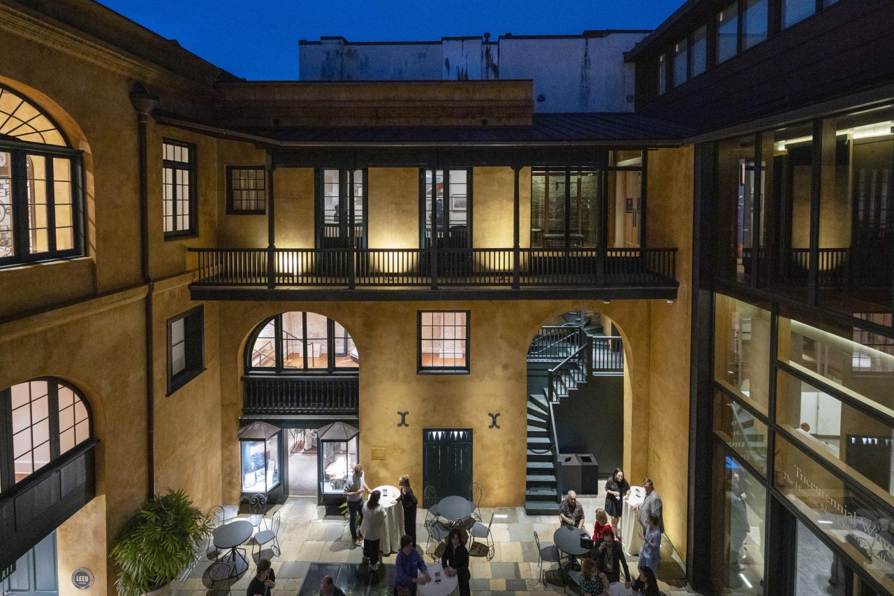 A courtyard at night features an elegant building with large windows and warm lighting. A staircase leads to the upper floor. People gather around tables below, enjoying the cozy ambiance.