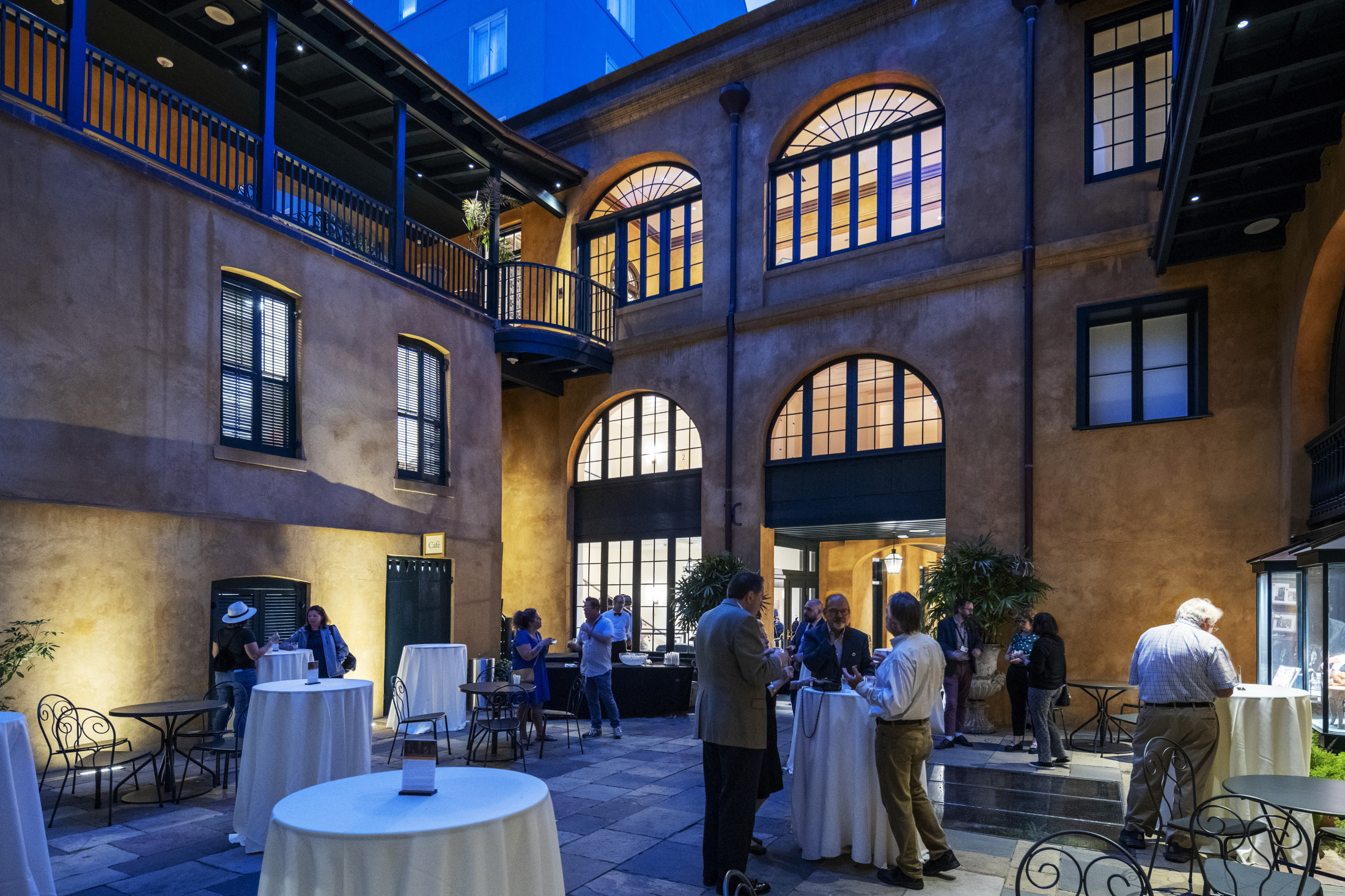 People gather in a well-lit, elegant courtyard with arched windows, tables, and chairs during an evening event. The setting includes historic architecture and a serene atmosphere.