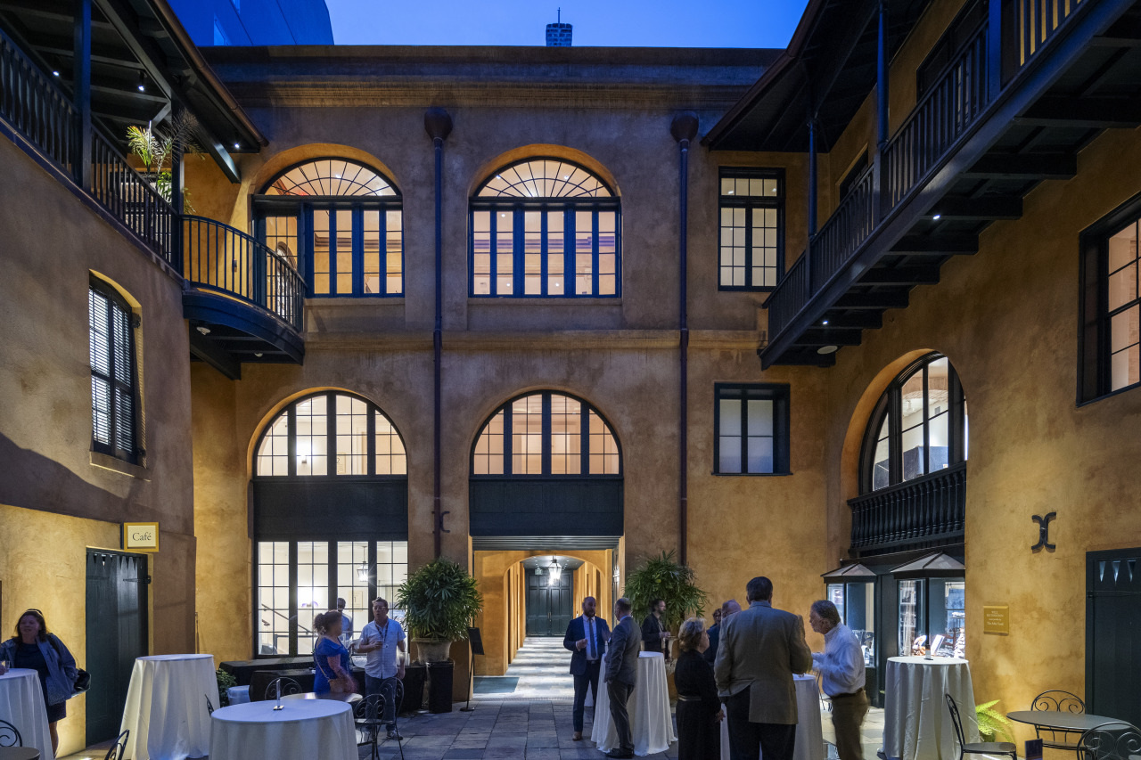 A group of people mingling in a rustic courtyard with arched windows and warm lighting. Tables are set with white tablecloths, and the ambient evening sky complements the cozy, historic architecture.