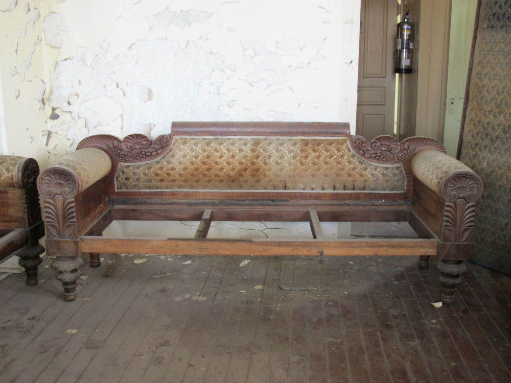 An antique wooden sofa with ornate carved details and faded upholstery sits in a room with peeling paint on the walls and a wooden floor. The sofa frame is exposed, missing its seat cushion.