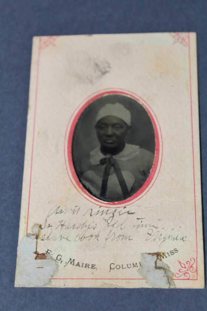 A vintage photograph of a person wearing a white hat and striped clothing, mounted on a card with handwritten text. The edges of the card are worn and discolored, with a decorative border and text indicating E.G. Maire, Columbus, Miss.